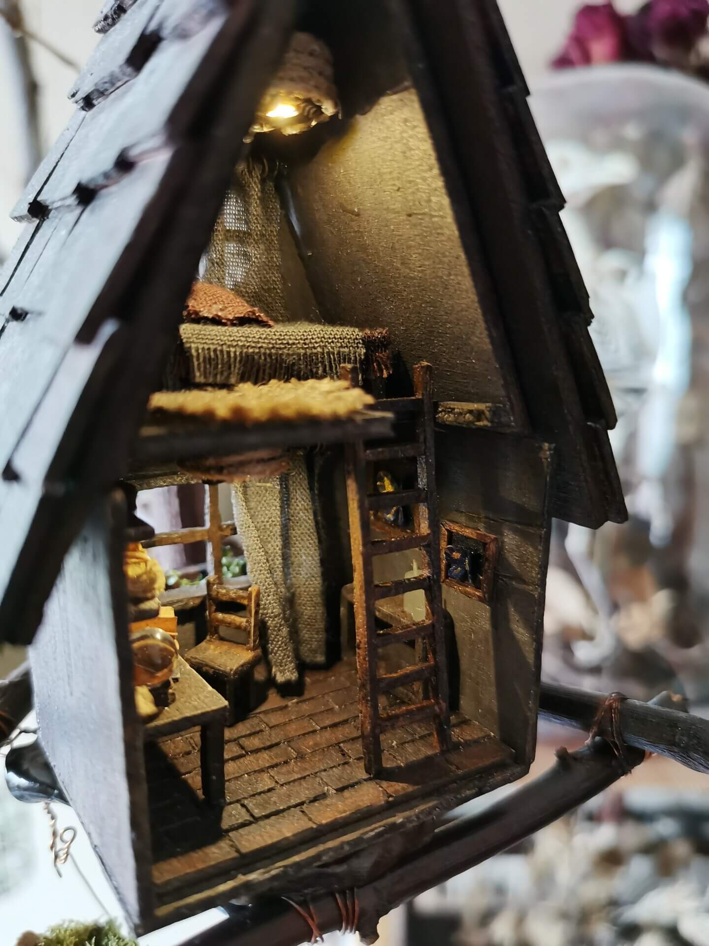 This miniature wooden treehouse exudes a whimsical, fairy-tale charm. It features a cozy loft bed under a slanted roof, accessible by a rustic wooden ladder. Warm lighting illuminates the intricate interior details, including a tiny desk, books, and woven fabrics, showcasing the delicate craftsmanship and magical ambiance of this enchanting creation.