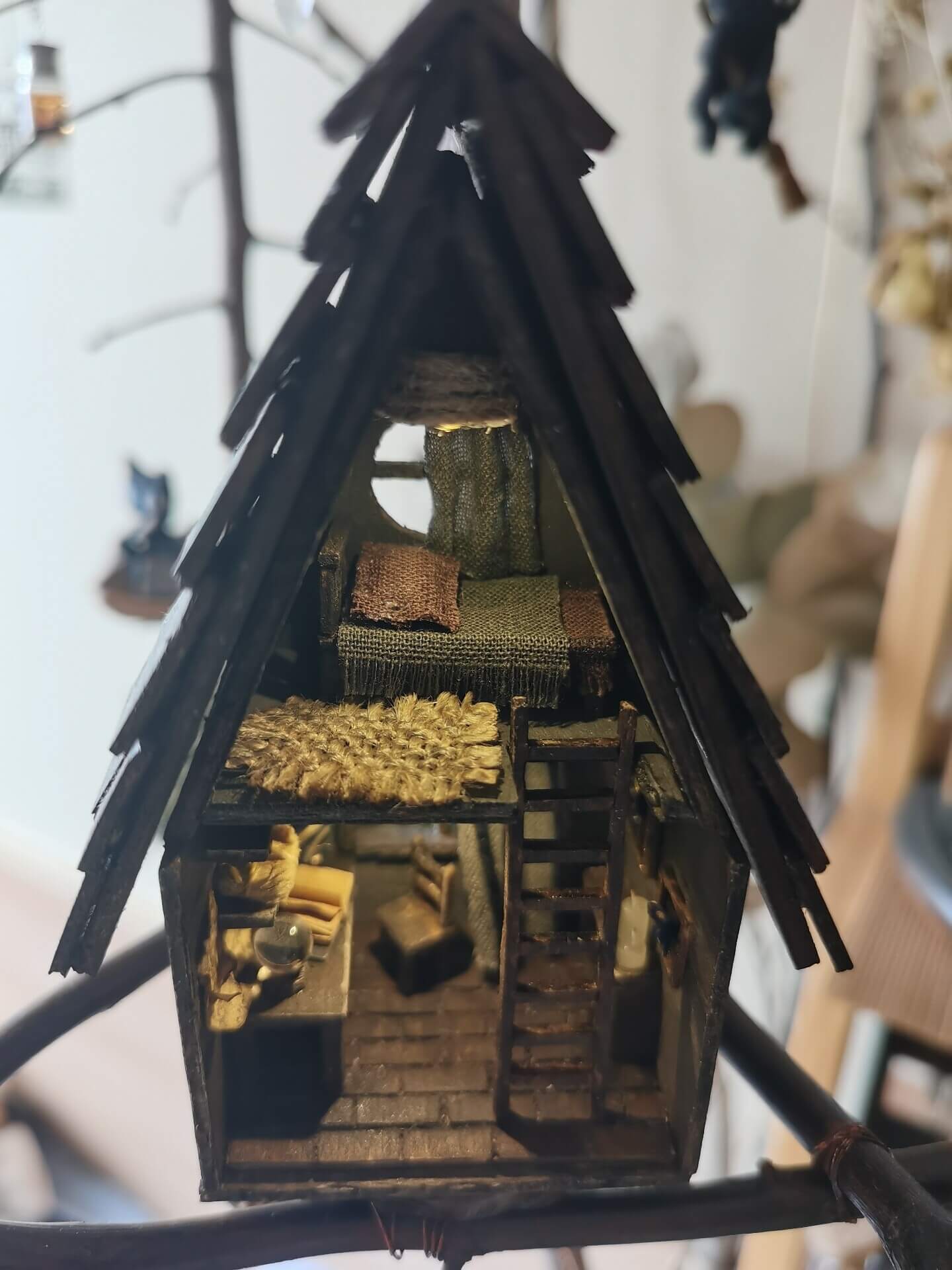 This miniature wooden treehouse exudes a whimsical, fairy-tale charm. It features a cozy loft bed under a slanted roof, accessible by a rustic wooden ladder. Warm lighting illuminates the intricate interior details, including a tiny desk, books, and woven fabrics, showcasing the delicate craftsmanship and magical ambiance of this enchanting creation.