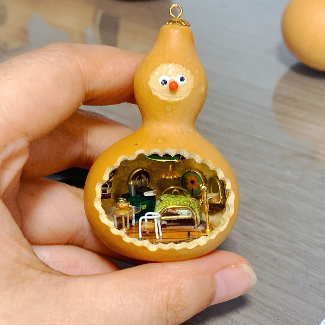 This adorable handcrafted gourd features a miniature bedroom tucked inside, complete with a cozy bed, a bedside table, and warm lighting. The intricate details, from the textured bedspread to the tiny furniture, create a whimsical and inviting space. Topped with a cheerful face, this gourd transforms into a charming piece of art that feels alive with personality and creativity.