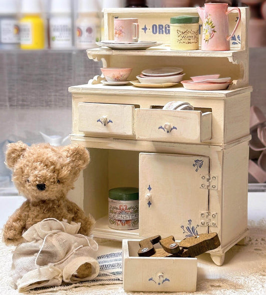 Add a touch of vintage charm to your dollhouse with this Miniature Vintage Kitchen Cabinet with Drawers and Shelves Kit. This beautifully crafted cabinet features multiple drawers and an open shelf, perfect for displaying or storing miniature kitchen items like plates, cups, and utensils. The charming floral decorations and soft yellow finish make this piece a perfect addition to any dollhouse kitchen, dining room, or living room.