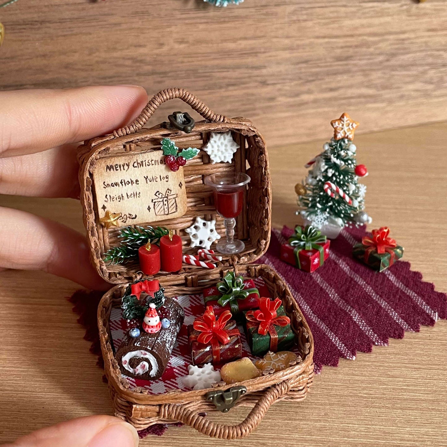 This festive miniature basket captures the magic of Christmas with incredible detail. Inside, you'll find tiny wrapped gifts, a cake, red candles, and a glass of wine, all arranged on a checkered cloth. The scene is complete with a mini Christmas tree and joyful holiday decorations, creating a cozy and enchanting holiday atmosphere.