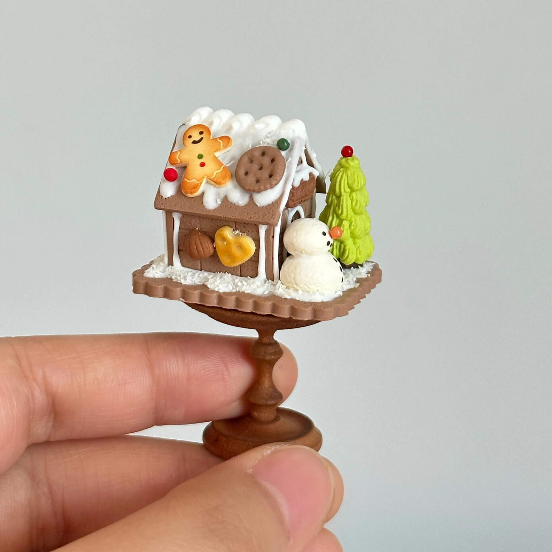 This delightful miniature diorama features a charming gingerbread house, beautifully crafted with intricate details that bring a festive winter wonderland to life. The house is adorned with snow-like frosting, colorful candies, and gingerbread cookies, giving it a cozy, holiday spirit. A tiny snowman stands beside the house, while a small green Christmas tree adds to the seasonal charm. The delicate snow and icing on the roof create a perfect winter scene, all set on a lovely circular wooden base.