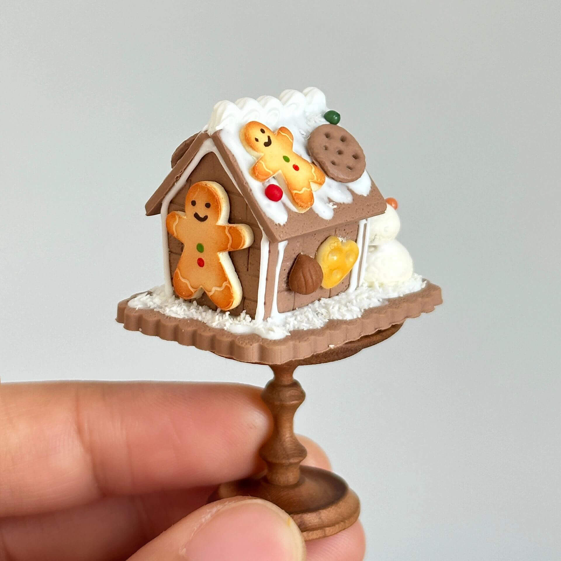 This delightful miniature diorama features a charming gingerbread house, beautifully crafted with intricate details that bring a festive winter wonderland to life. The house is adorned with snow-like frosting, colorful candies, and gingerbread cookies, giving it a cozy, holiday spirit. A tiny snowman stands beside the house, while a small green Christmas tree adds to the seasonal charm. The delicate snow and icing on the roof create a perfect winter scene, all set on a lovely circular wooden base.