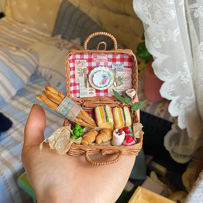 This charming miniature picnic basket brings the essence of a French picnic to life. With a red checkered lining, it features mini baguettes, sandwiches, croissants, peas, and strawberries. A delicate porcelain plate and cutlery add a touch of elegance, while intricate paper details and a tiny rose complete the idyllic scene. Perfectly crafted, it's a delightful escape into the art of miniatures!