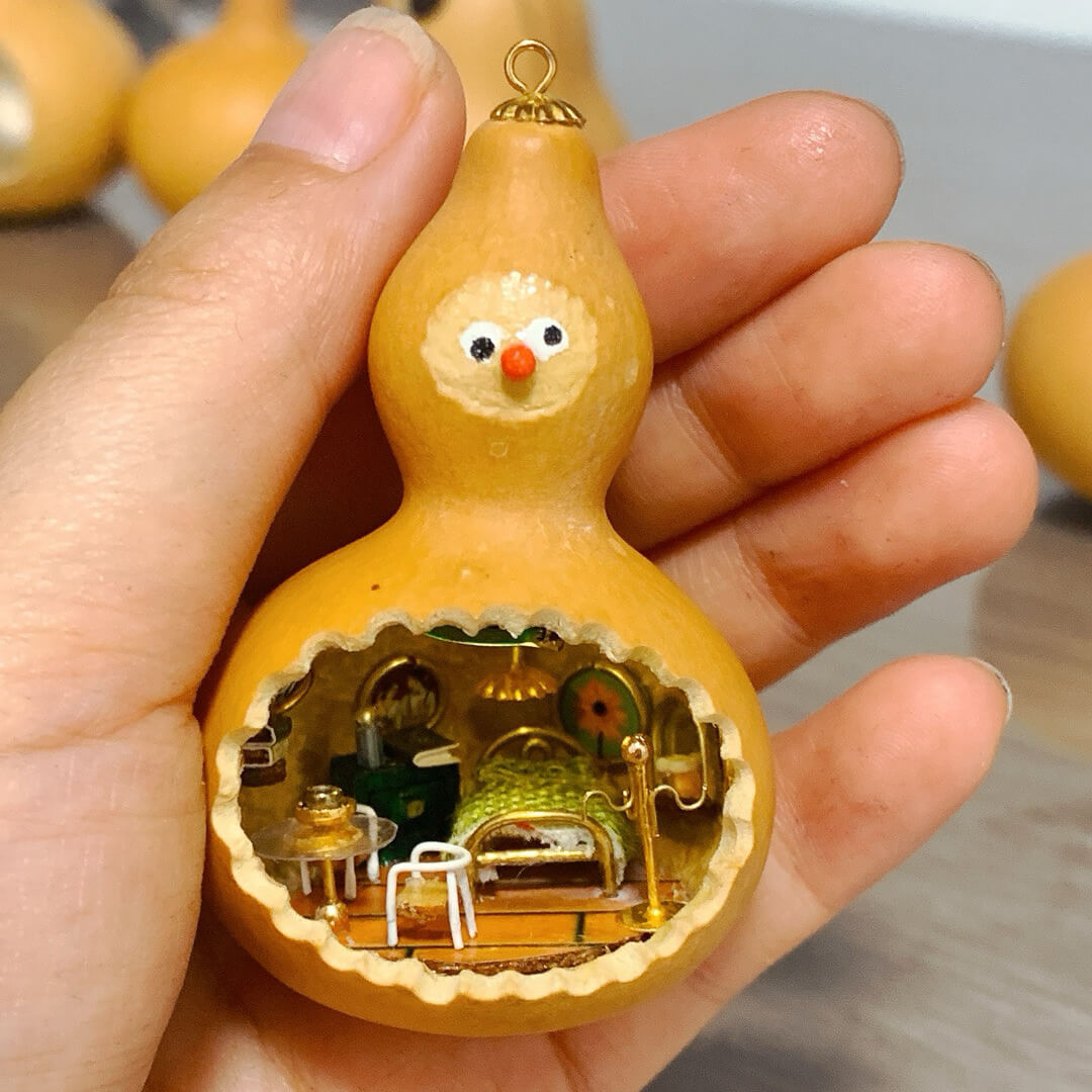 This adorable handcrafted gourd features a miniature bedroom tucked inside, complete with a cozy bed, a bedside table, and warm lighting. The intricate details, from the textured bedspread to the tiny furniture, create a whimsical and inviting space. Topped with a cheerful face, this gourd transforms into a charming piece of art that feels alive with personality and creativity.