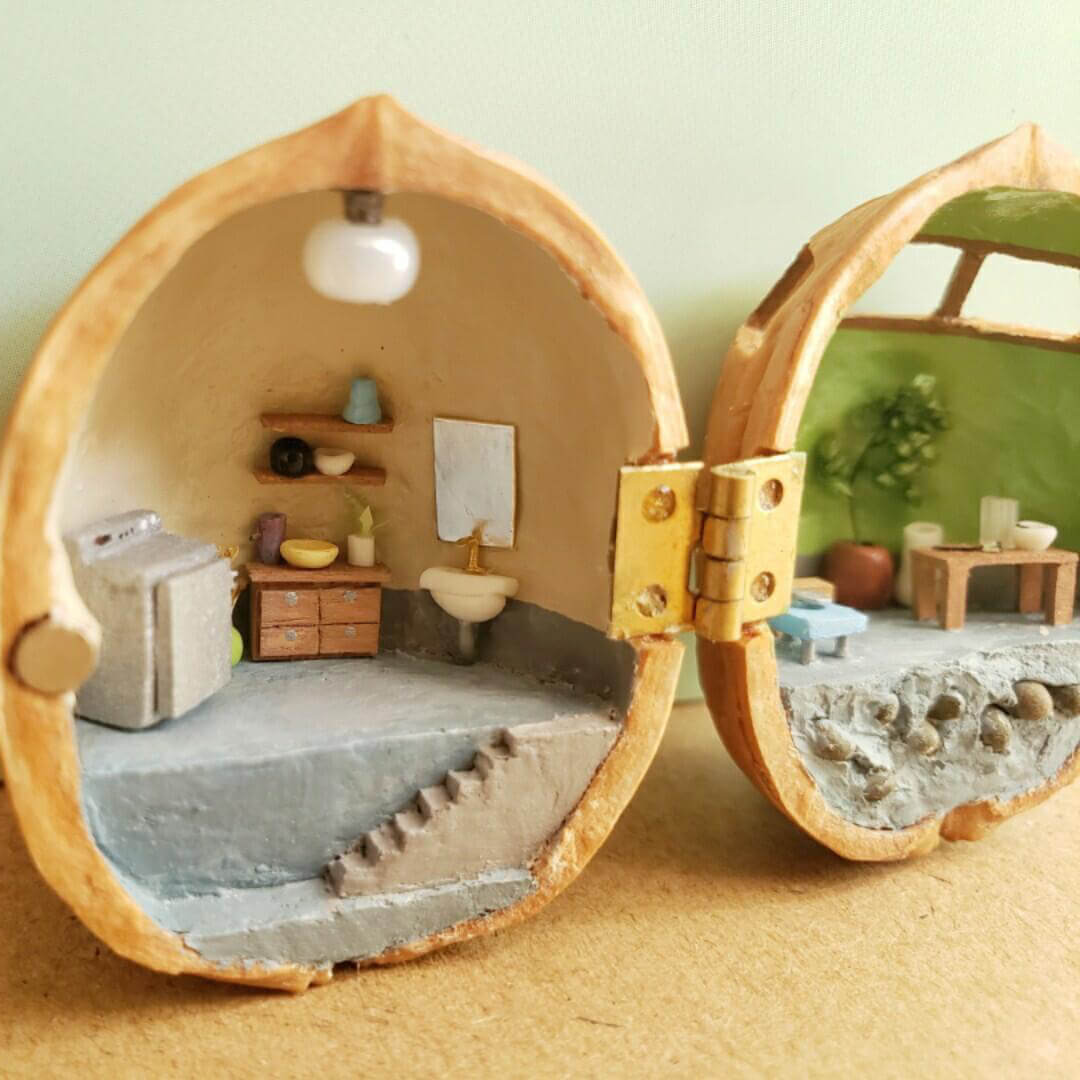 This intricate miniature scene transforms a walnut shell into a charming ceramic studio. On one side, the studio workspace is equipped with a chest of drawers, and shelves holding tiny pottery tools and supplies. The other side features a serene crafting area with a green backdrop, a table, and display shelves showcasing finished ceramic pieces.