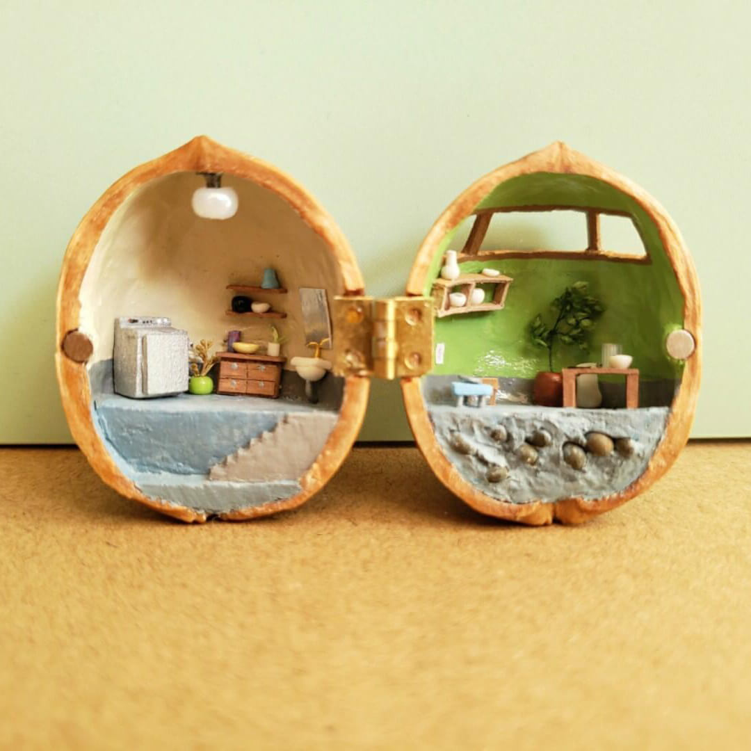 This intricate miniature scene transforms a walnut shell into a charming ceramic studio. On one side, the studio workspace is equipped with a chest of drawers, and shelves holding tiny pottery tools and supplies. The other side features a serene crafting area with a green backdrop, a table, and display shelves showcasing finished ceramic pieces.