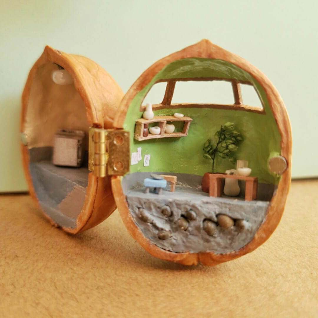 This intricate miniature scene transforms a walnut shell into a charming ceramic studio. On one side, the studio workspace is equipped with a chest of drawers, and shelves holding tiny pottery tools and supplies. The other side features a serene crafting area with a green backdrop, a table, and display shelves showcasing finished ceramic pieces.