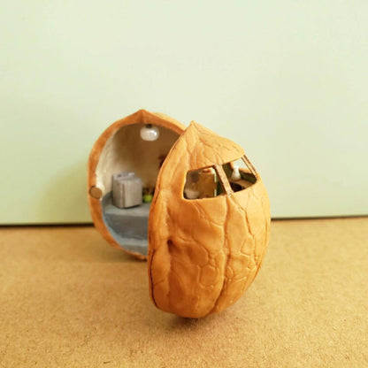 This intricate miniature scene transforms a walnut shell into a charming ceramic studio. On one side, the studio workspace is equipped with a chest of drawers, and shelves holding tiny pottery tools and supplies. The other side features a serene crafting area with a green backdrop, a table, and display shelves showcasing finished ceramic pieces.