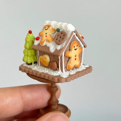 This delightful miniature diorama features a charming gingerbread house, beautifully crafted with intricate details that bring a festive winter wonderland to life. The house is adorned with snow-like frosting, colorful candies, and gingerbread cookies, giving it a cozy, holiday spirit. A tiny snowman stands beside the house, while a small green Christmas tree adds to the seasonal charm. The delicate snow and icing on the roof create a perfect winter scene, all set on a lovely circular wooden base.