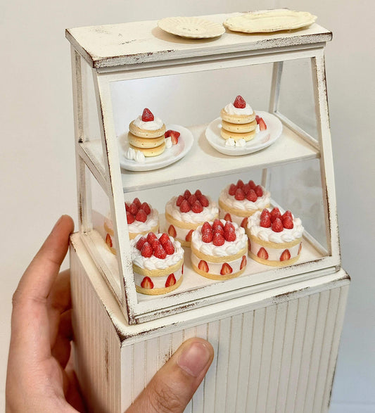 This charming miniature dessert display case is a sweet-tooth's dream brought to life. Featuring perfectly detailed strawberry shortcakes and pancake, each creation looks as delectable as the real thing. The rustic white display cabinet adds a cozy bakery vibe, enhancing the charm of these handcrafted treats. Every element, from the whipped cream to the vibrant strawberries, showcases the meticulous artistry and attention to detail.