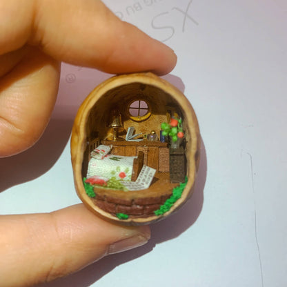 This enchanting walnut shell reveals a miniature countryside bedroom. With a neatly made bed adorned with a floral blanket, a wooden desk holding tiny books and objects, and a glowing lamp, the cozy interior is surrounded by green foliage. The tiny window completes the serene, homey atmosphere, making this piece a delightful fusion of craftsmanship and charm.