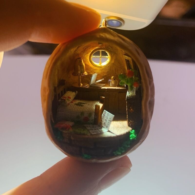 This enchanting walnut shell reveals a miniature countryside bedroom. With a neatly made bed adorned with a floral blanket, a wooden desk holding tiny books and objects, and a glowing lamp, the cozy interior is surrounded by green foliage. The tiny window completes the serene, homey atmosphere, making this piece a delightful fusion of craftsmanship and charm.