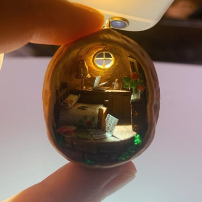 This enchanting walnut shell reveals a miniature countryside bedroom. With a neatly made bed adorned with a floral blanket, a wooden desk holding tiny books and objects, and a glowing lamp, the cozy interior is surrounded by green foliage. The tiny window completes the serene, homey atmosphere, making this piece a delightful fusion of craftsmanship and charm.