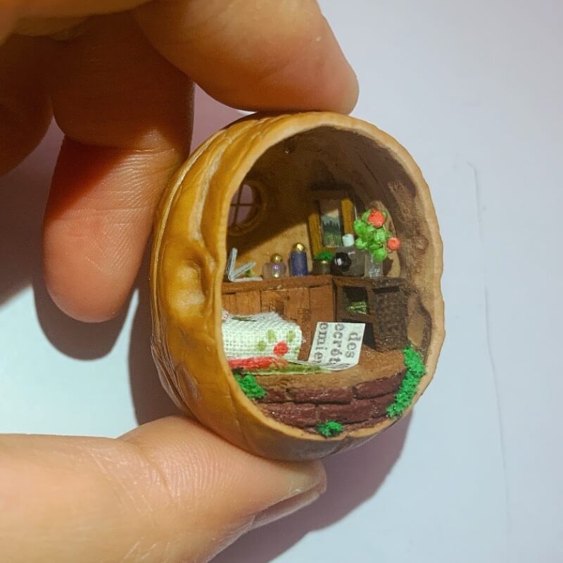 This enchanting walnut shell reveals a miniature countryside bedroom. With a neatly made bed adorned with a floral blanket, a wooden desk holding tiny books and objects, and a glowing lamp, the cozy interior is surrounded by green foliage. The tiny window completes the serene, homey atmosphere, making this piece a delightful fusion of craftsmanship and charm.