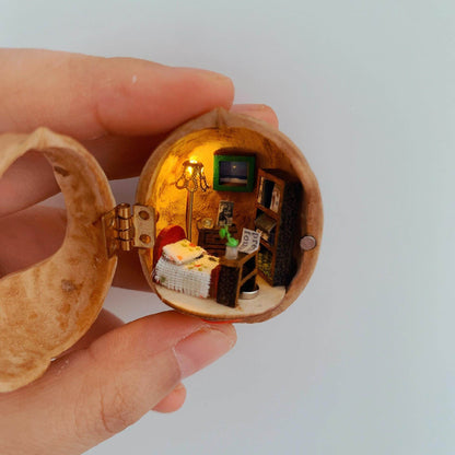 This walnut shell contains a charmingly detailed miniature bedroom. It features a cozy bed with a textured blanket, a tiny bookshelf stacked with books, a warm floor lamp, and a tiny potted plant. The soft lighting and carefully crafted details create a peaceful and intimate ambiance, perfect for a quiet escape.