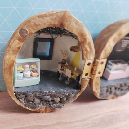 This is a charming miniature dessert shop tucked inside a walnut shell, radiating a whimsical, fairytale-like charm. On one side, there’s a dessert display case filled with tiny cakes and pastries, intricately detailed. On the other side, a cozy dining area features small tables, chairs, and a soft glowing lamp, creating a warm and inviting atmosphere.