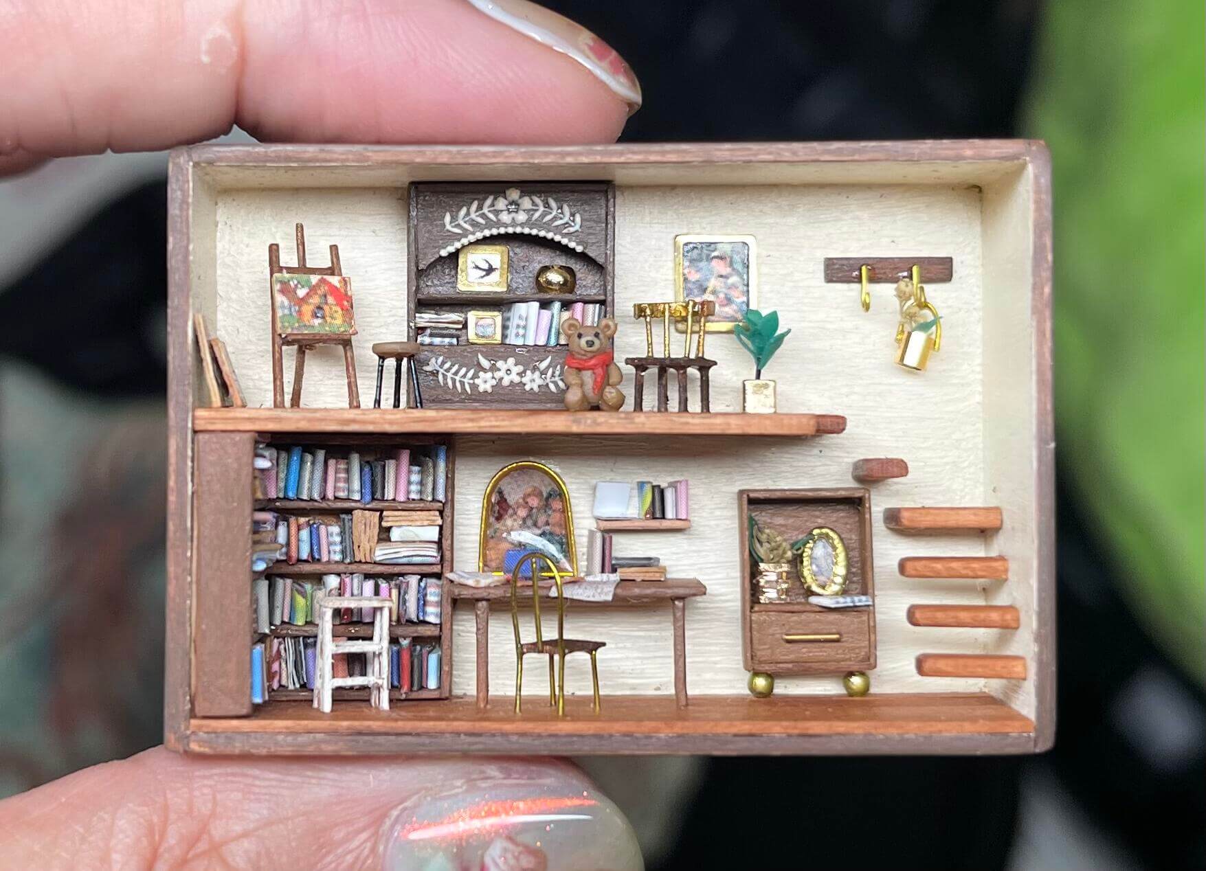 This miniature scene captures the charm of a cozy home library and art studio, complete with tiny bookshelves, a delicate art easel displaying a painting, a comfortable reading nook, and intricate decor details. Every element, from the tiny teddy bear on the shelf to the potted plant and framed artwork, brings warmth and personality to this tiny creative retreat.