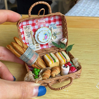 This charming miniature picnic basket brings the essence of a French picnic to life. With a red checkered lining, it features mini baguettes, sandwiches, croissants, peas, and strawberries. A delicate porcelain plate and cutlery add a touch of elegance, while intricate paper details and a tiny rose complete the idyllic scene. Perfectly crafted, it's a delightful escape into the art of miniatures!