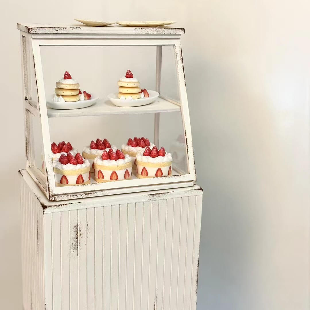 This charming miniature dessert display case is a sweet-tooth's dream brought to life. Featuring perfectly detailed strawberry shortcakes and pancake, each creation looks as delectable as the real thing. The rustic white display cabinet adds a cozy bakery vibe, enhancing the charm of these handcrafted treats. Every element, from the whipped cream to the vibrant strawberries, showcases the meticulous artistry and attention to detail.