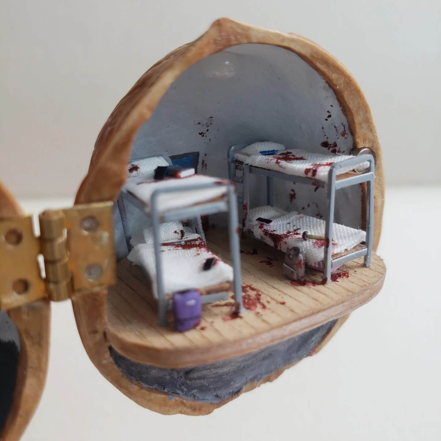 A hauntingly detailed miniature diorama set inside a walnut shell, capturing an eerie and unsettling atmosphere. One side depicts a blood-streaked door labeled "302", with scattered papers adding to the mystery. The other side reveals a grim dormitory-style room with bunk beds, bloodstains, and abandoned belongings, hinting at a dark story. The meticulous craftsmanship and chilling details evoke a sense of suspense and curiosity, making this piece a unique and thought-provoking work of art.