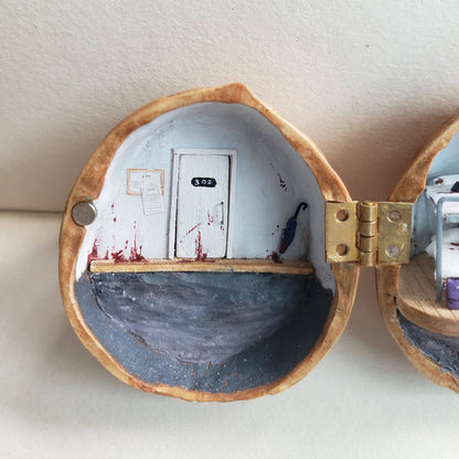 A hauntingly detailed miniature diorama set inside a walnut shell, capturing an eerie and unsettling atmosphere. One side depicts a blood-streaked door labeled "302", with scattered papers adding to the mystery. The other side reveals a grim dormitory-style room with bunk beds, bloodstains, and abandoned belongings, hinting at a dark story. The meticulous craftsmanship and chilling details evoke a sense of suspense and curiosity, making this piece a unique and thought-provoking work of art.