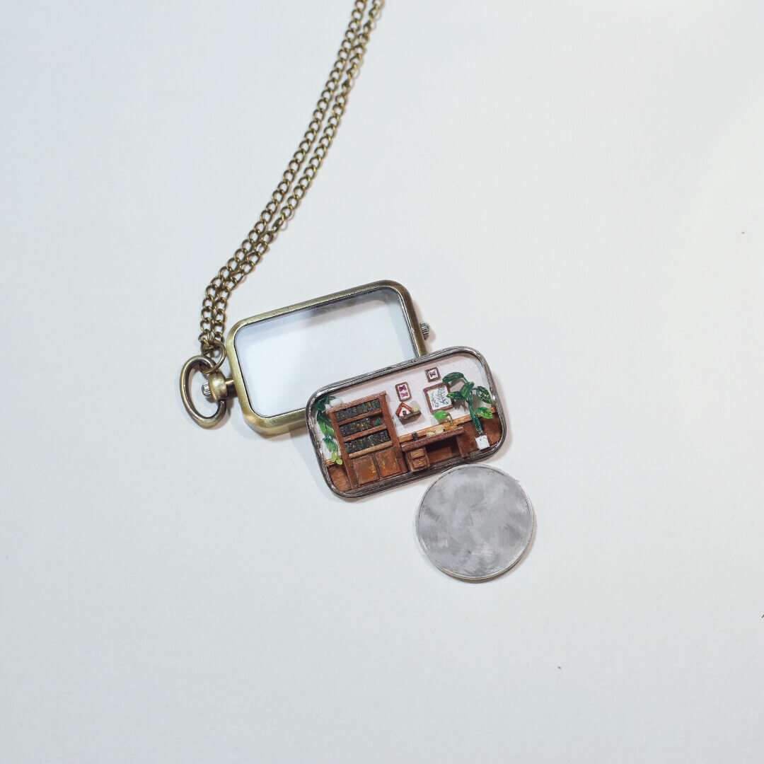 This is a delicate miniature pendant featuring a tiny study room. Inside the pendant’s frame lies a meticulously crafted scene, complete with a wooden desk, bookshelf, potted plants, and tiny decorations, evoking the warmth and charm of a cozy workspace. Perfect as both jewelry and an art piece, it combines creativity and craftsmanship in an enchanting way.