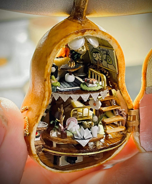 This intricate diorama captures a magical miniature home inside a gourd. The multi-level design showcases a cozy bedroom, a charming dining setup, and whimsical decorations. Adorned with delicate bunting, flowers, books, and candles, every detail exudes warmth and creativity. This enchanting craft is a tiny masterpiece that feels alive with stories waiting to unfold.