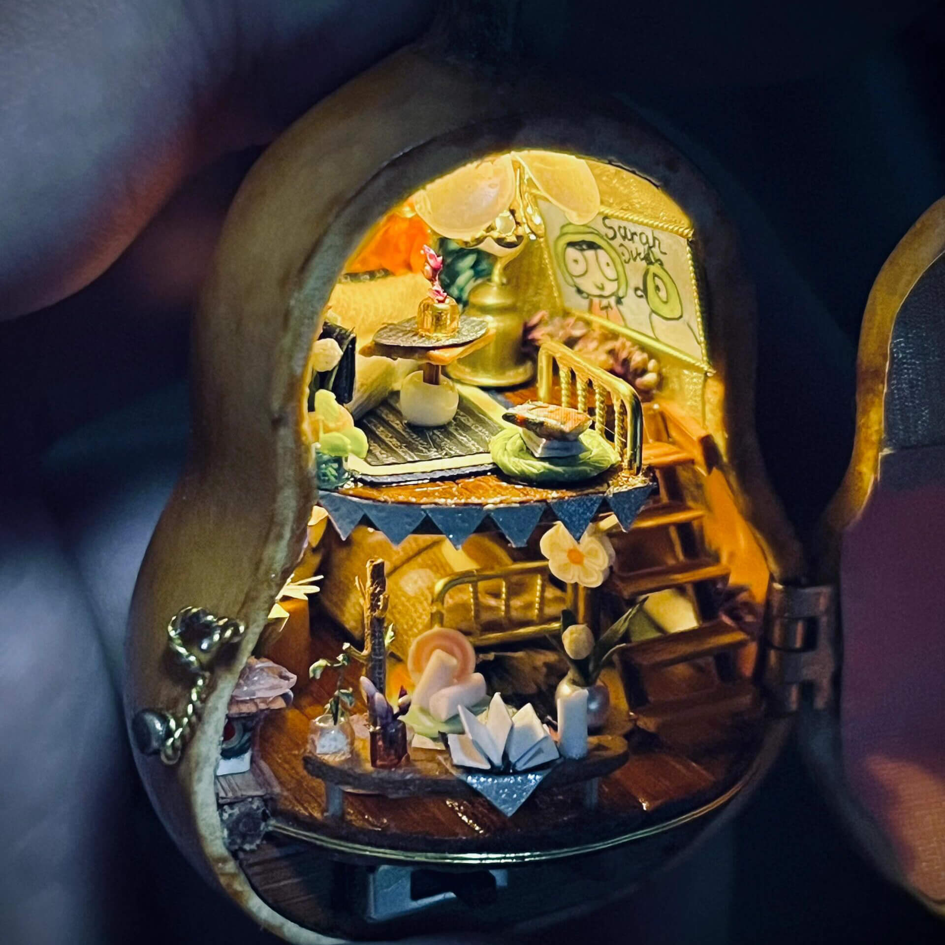 This intricate diorama captures a magical miniature home inside a gourd. The multi-level design showcases a cozy bedroom, a charming dining setup, and whimsical decorations. Adorned with delicate bunting, flowers, books, and candles, every detail exudes warmth and creativity. This enchanting craft is a tiny masterpiece that feels alive with stories waiting to unfold.